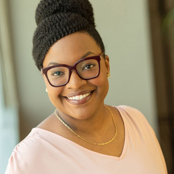 Portrait of Tonya Nelson smiling in thick-rimmed brown glasses, a pink shirt, and a thin gold chain necklace with matching earrings.