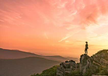 A woman stands silhouetted at the top of a mountain backlit by the setting sun.