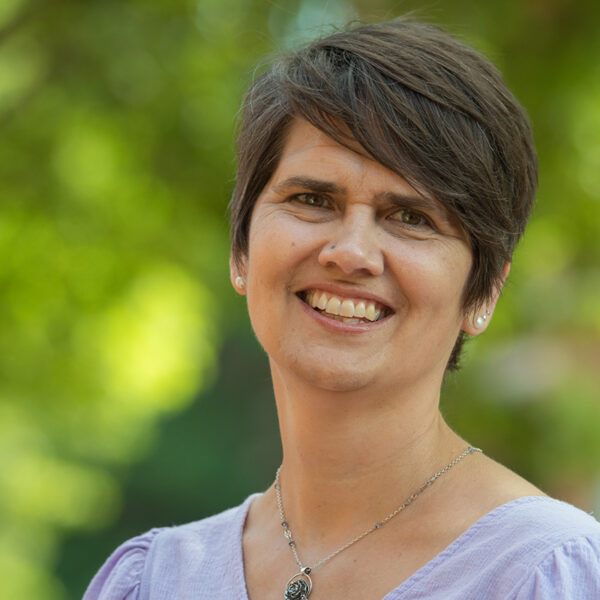 Portrait of Anya Shepelavey smiling in a lilac top and silver rose pendant necklace.